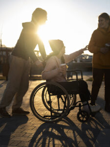 Grupo de três jovens amigos interagindo em um evento cultural ao ar livre durante o pôr do sol. Uma pessoa está sentada em uma cadeira de rodas e sorri enquanto alcança o braço de outra pessoa que está em pé ao lado. Um terceiro amigo empurra a cadeira de rodas.
