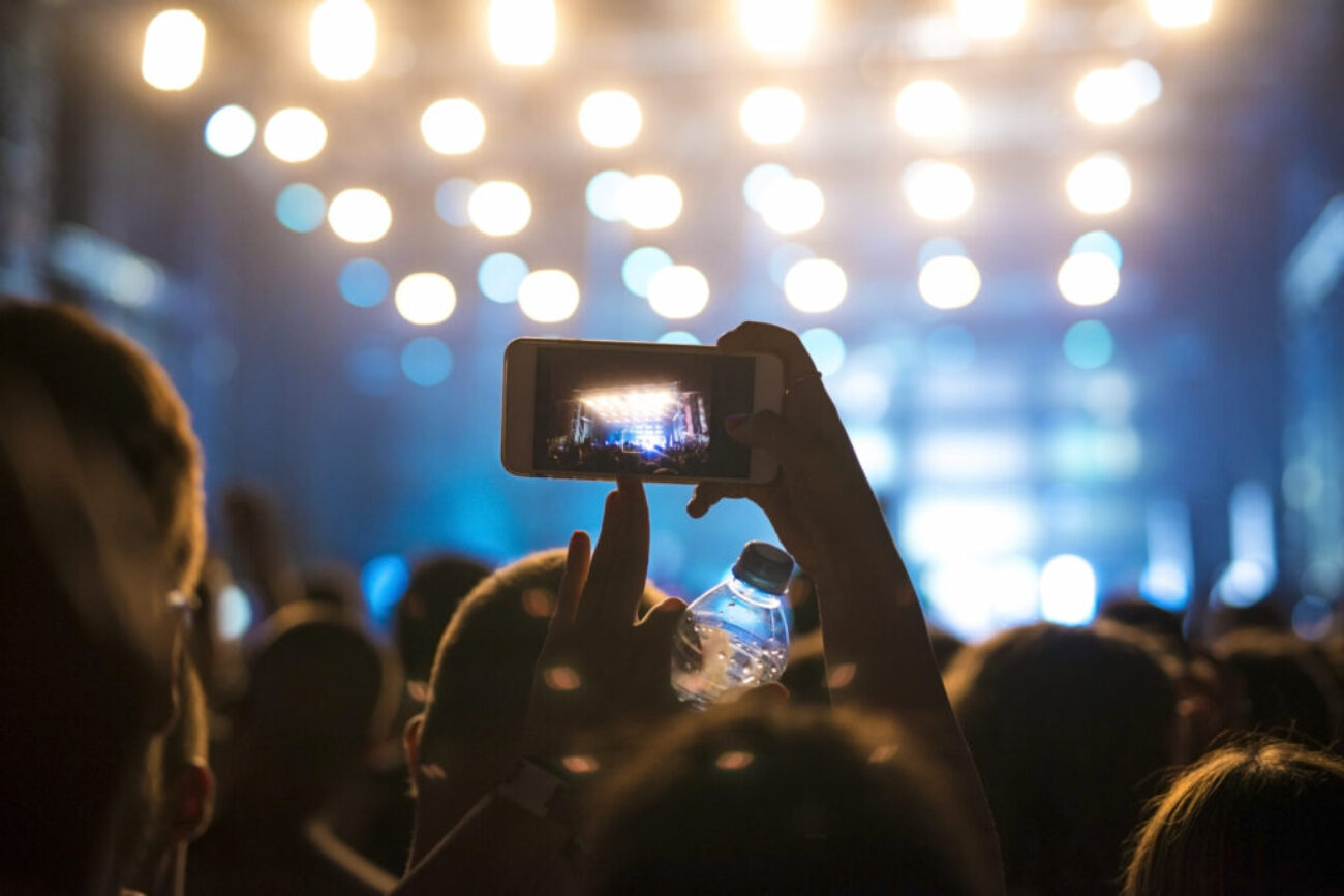 Multidão animada assistindo a um show ou festival à noite, com uma pessoa segurando um celular para gravar a apresentação no palco iluminado.