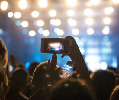 Multidão animada assistindo a um show ou festival à noite, com uma pessoa segurando um celular para gravar a apresentação no palco iluminado.