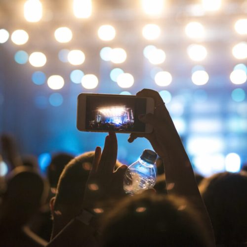 Multidão animada assistindo a um show ou festival à noite, com uma pessoa segurando um celular para gravar a apresentação no palco iluminado.