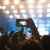 Multidão animada assistindo a um show ou festival à noite, com uma pessoa segurando um celular para gravar a apresentação no palco iluminado.